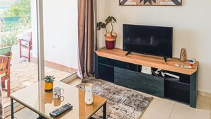 Una sala de estar con un televisor de pantalla plana sobre un soporte de madera, una planta en maceta, un control remoto y una taza de café sobre una mesa de vidrio.
