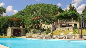 Una piscina amb gandules i una zona coberta, envoltada per arbres i edificis de pedra.