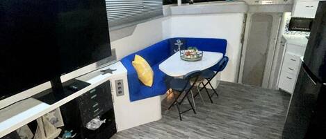 A compact living space with a flat-screen TV, a small dining table with blue cushions, and a kitchenette area.