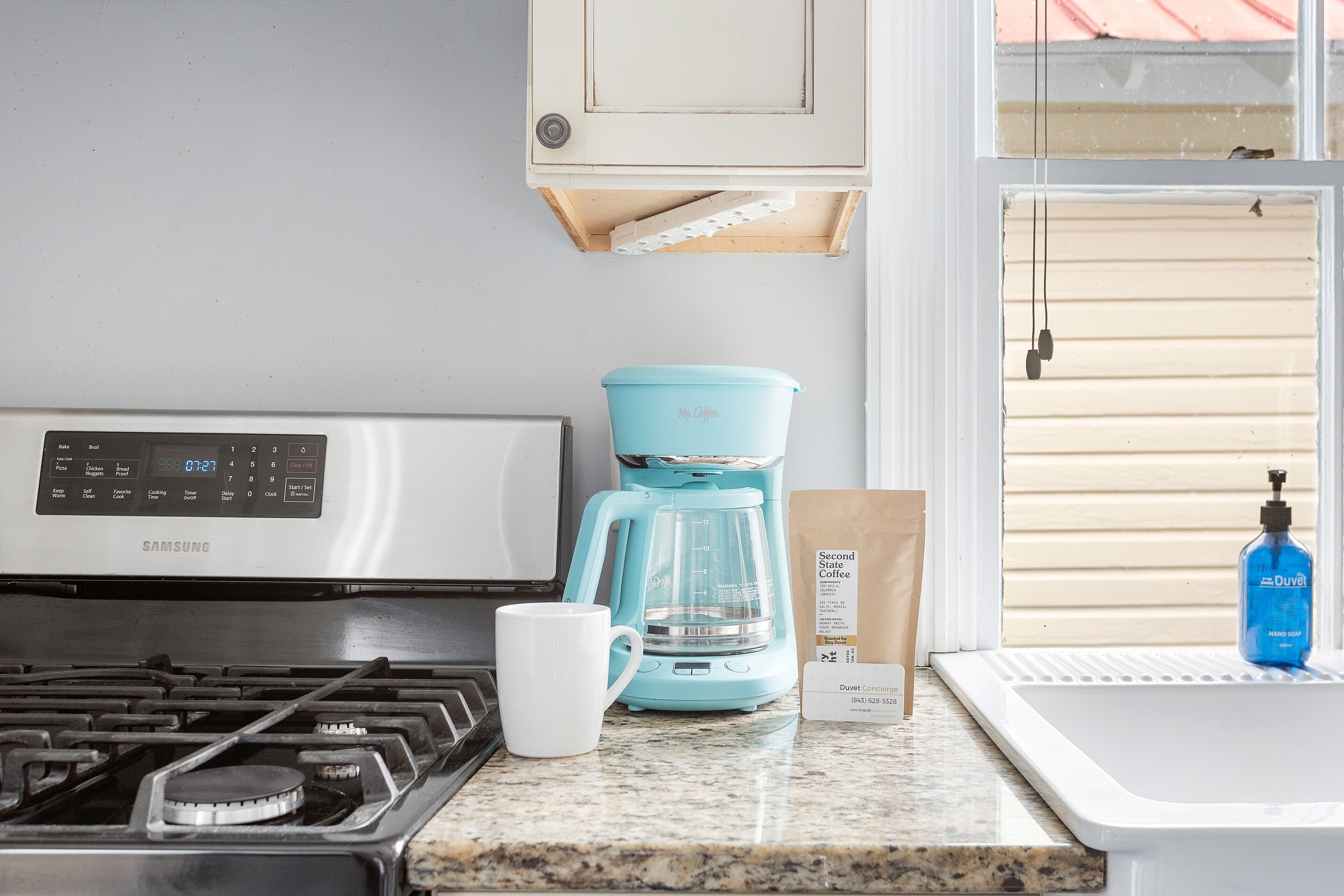 A kitchen with a stainless steel stove, a blue coffee maker, a white mug, and a Duet Concierge bag.