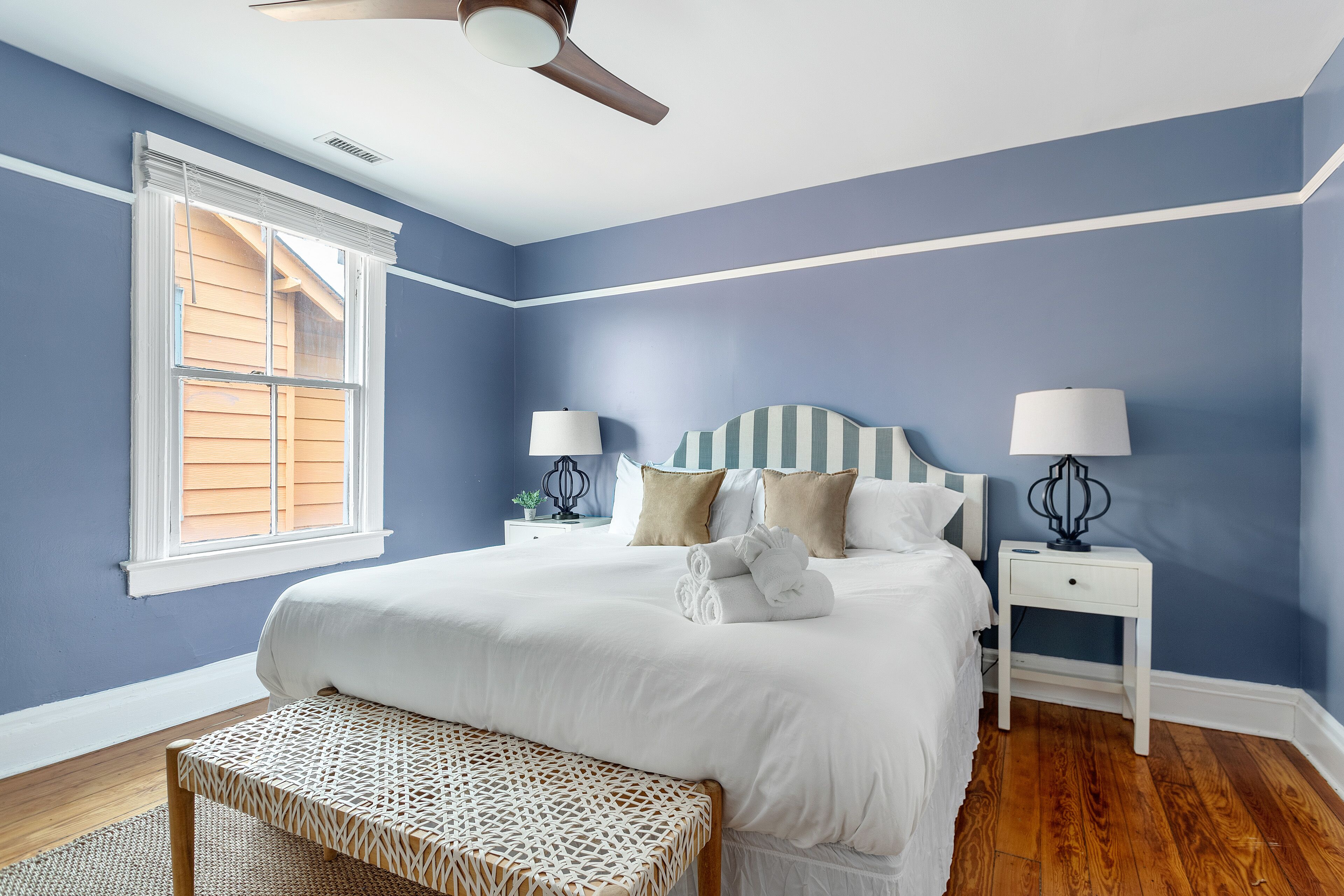A bedroom with a bed, two bedside tables, a bench, a ceiling fan, and a window with blinds.