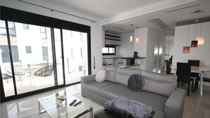 Una sala de estar moderna con un sofá gris, una mesa de centro de vidrio y un comedor con una mesa y sillas blancas.