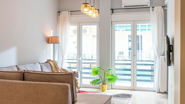 A living room with a sofa, a lamp, a plant, and a sliding glass door leading to a balcony. - 2 Bedroom & 2 Bathroom - 4mins Walk To Acropolis (Athina)