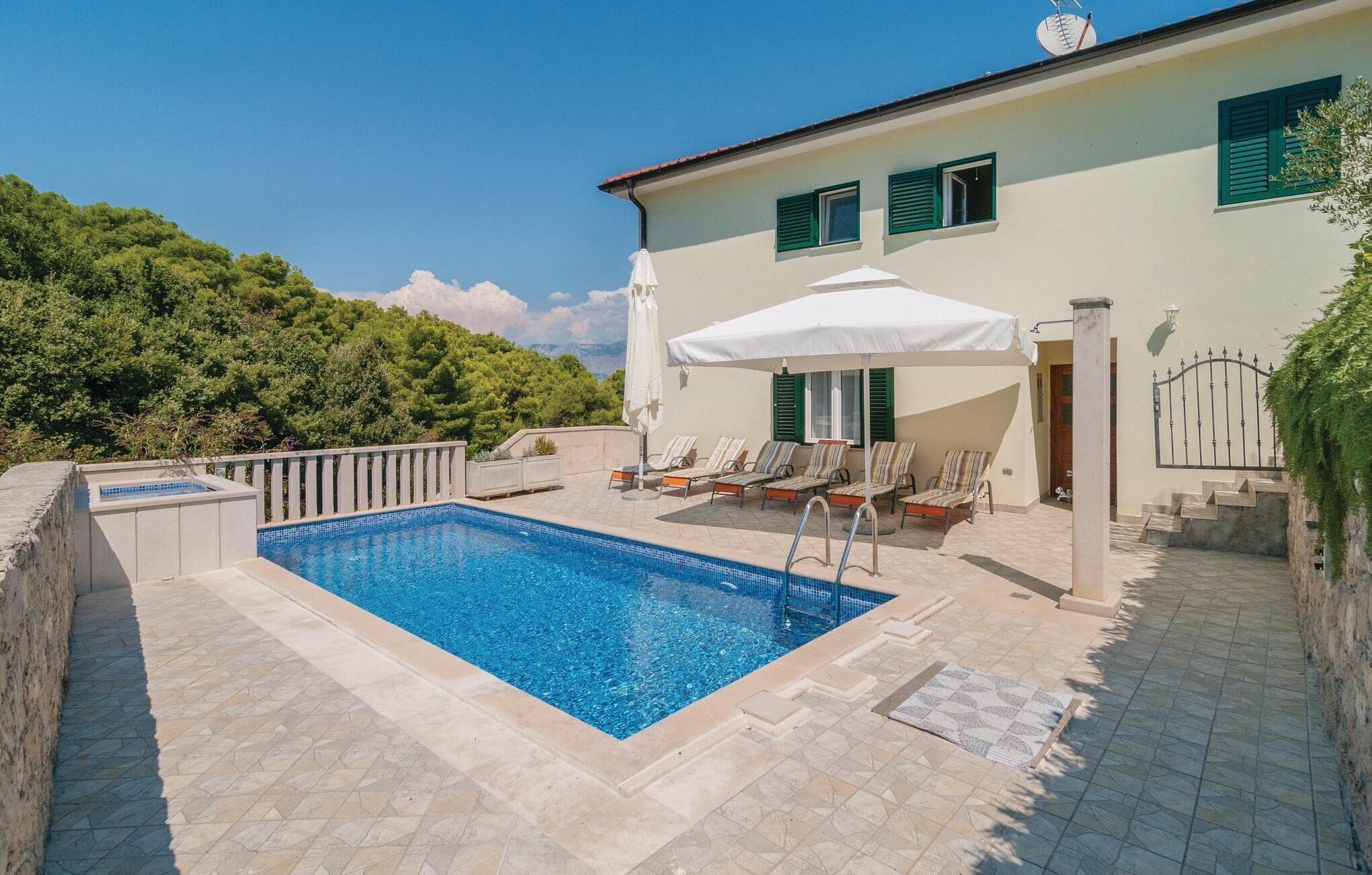 A swimming pool with lounge chairs and umbrellas, surrounded by a stone deck and a building with green shutters.