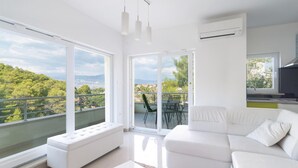 A modern living room with a white sofa, a white bench, and a dining area with a table and chairs. Large windows offer a view of a green landscape and buildings.