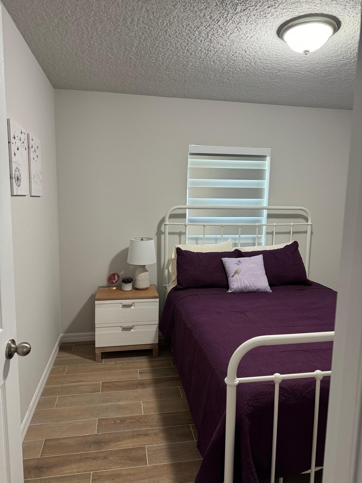 Una camera da letto con un letto, un comodino, una lampada, una finestra con tende e una porta.