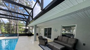 A covered outdoor pool area with a grill, wicker furniture, and a view of trees.