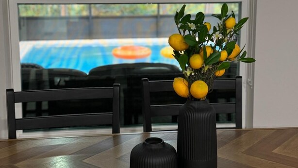 A dining area with a table and chairs, a vase with lemons, and a view through a window.