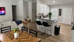 A modern kitchen with white cabinets, a black countertop, and a bar area. There is a dining table with chairs and a TV mounted on the wall.