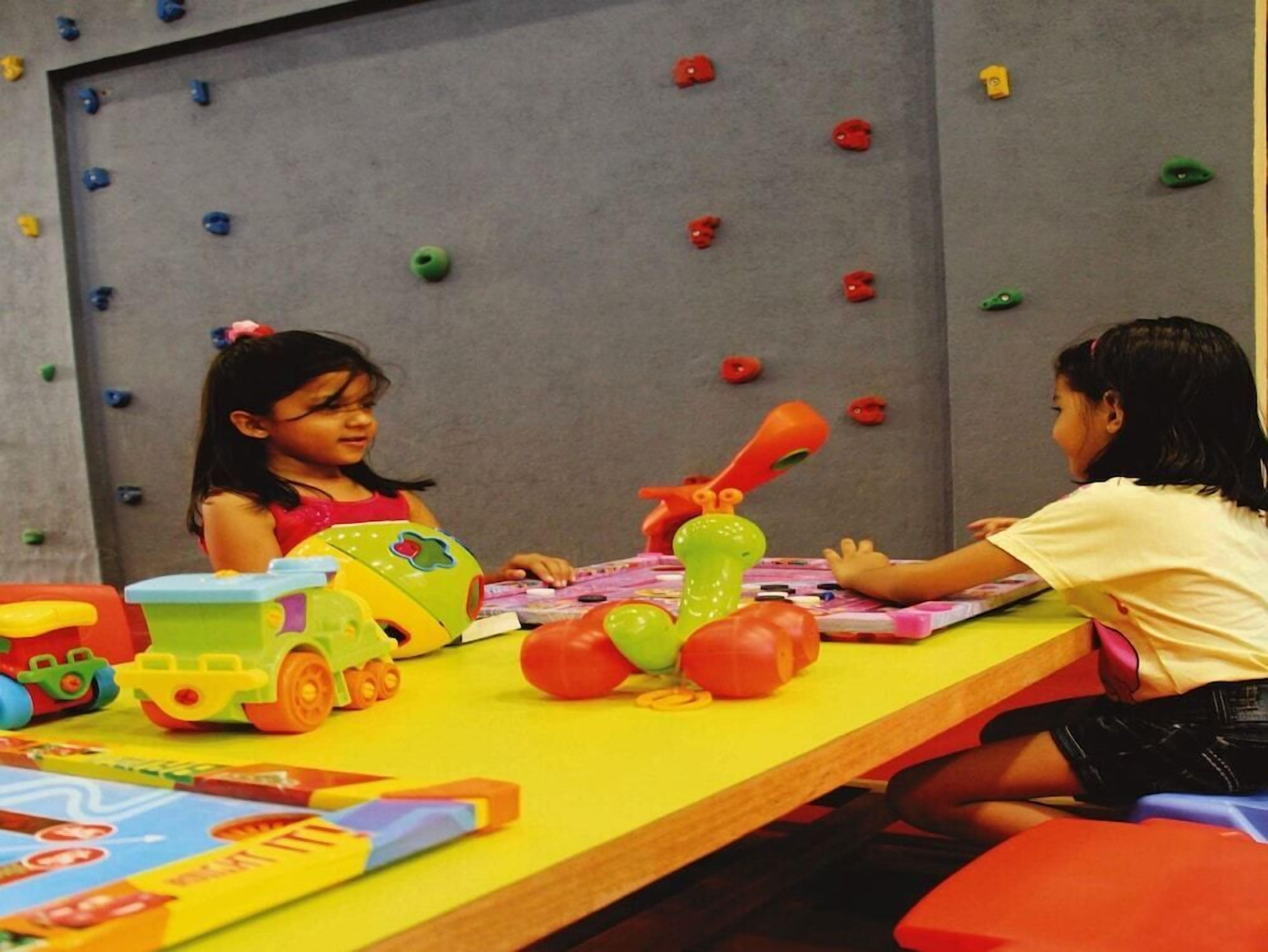 Duas crianças brincando com brinquedos a uma mesa em um quarto com uma parede de escalada ao fundo.