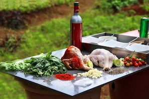 A table with raw chicken, various herbs, spices, and a bottle of sauce, set outdoors with a grassy background. - Oxford Golf Resort (Pune)