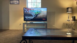 Un soggiorno con una televisione che mostra 'Car Masters Rust to Riches' su Netflix, una finestra con persiane, un cartello con la scritta 'Love' e una lampada su un tavolino.