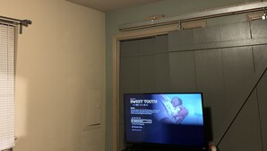Una stanza con ventilatore a soffitto, televisore su supporto e finestra con persiane.