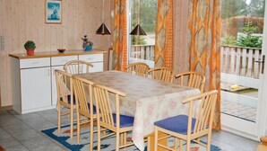 A dining area with a wooden table and chairs, a kitchen counter with cabinets, and a view of a garden through a window with patterned curtains. - Stunning home in Middelfart with sauna (Middelfart)
