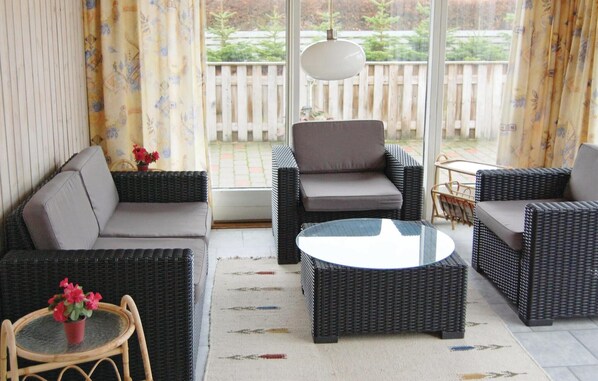 A sunlit patio with wicker furniture, a glass table, and a view of a wooden fence and greenery.
