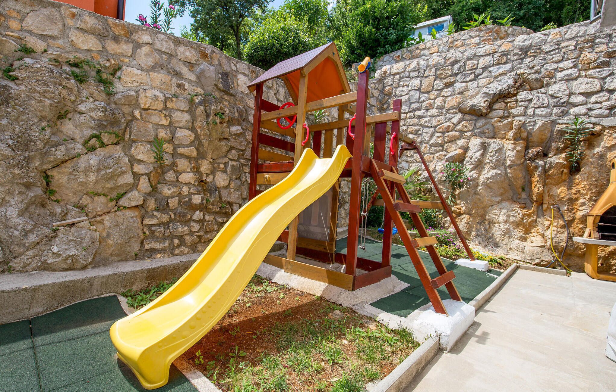 A playground with a yellow slide and a climbing structure, set against a stone wall and surrounded by greenery.