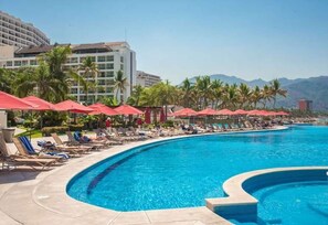 A resort pool area with sun loungers and umbrellas, surrounded by palm trees and a large hotel building.