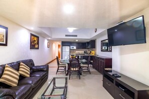 A modern living room with a leather sofa, a glass coffee table, a dining area with a table and chairs, a kitchen with dark cabinets, and a flat-screen TV mounted on the wall.