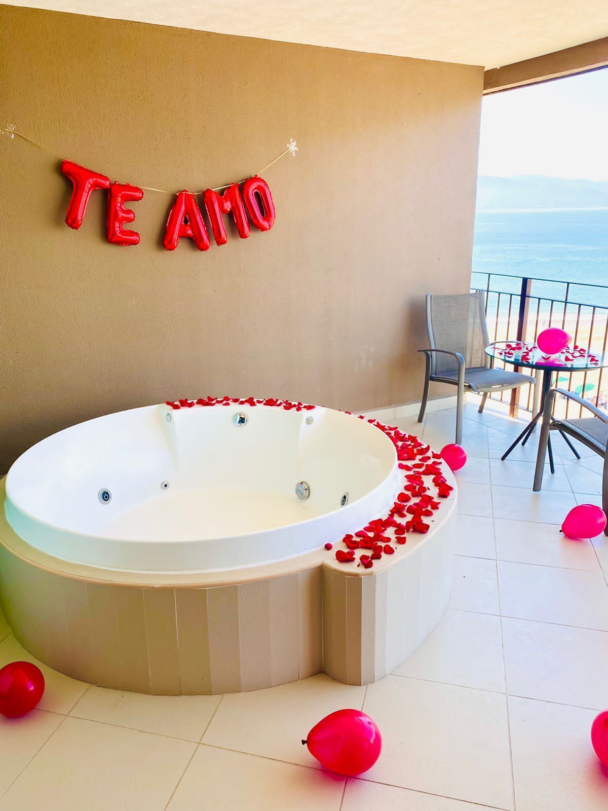 Un bain à remous décoré de pétales de rose et de ballons, une chaise et une table offrant une vue sur l’océan.
