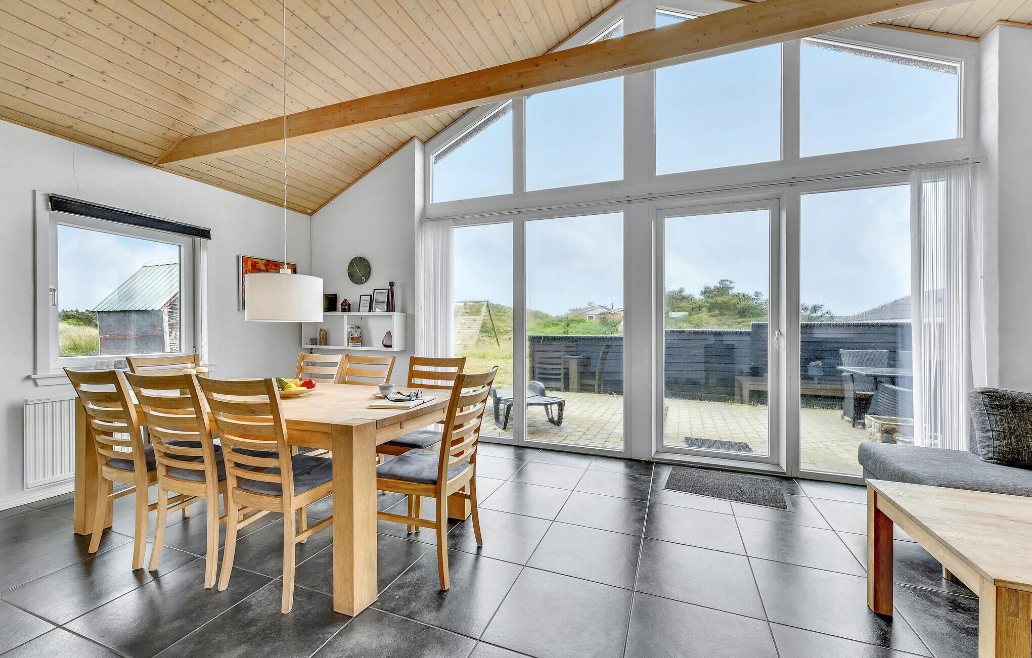 A dining area with a wooden table and chairs, large windows, and a view of the outdoors.