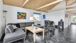 Una sala de estar moderna con un sofá seccional gris, una mesa de centro de madera y un sillón de cuero negro. Se observan pinturas abstractas en las paredes y un ventanal que proporciona luz natural.