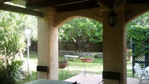 Un espace extérieur aménagé en terrasse, avec une table et des chaises, un luminaire suspendu, et une vue sur un jardin avec un arbre et un mur en pierre.