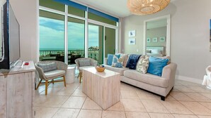 A spacious living room with a large window offering a view of the ocean, a comfortable sofa set, a wooden coffee table, and a wicker chandelier. - Sapphire 505 - By GISO Vacation Rentals (South Padre Island)
