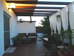 A covered patio area with two chairs and a table, surrounded by potted plants and a hanging plant.