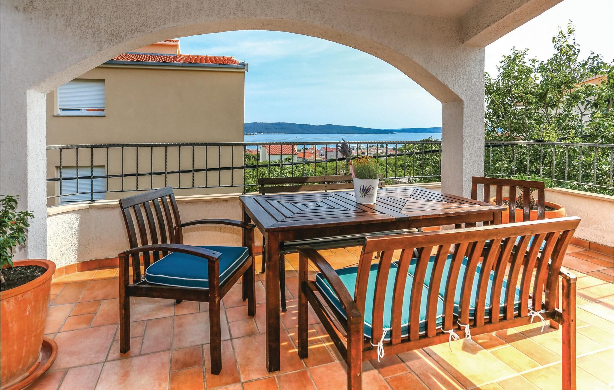 A terrace with a wooden table and chairs, a potted plant, and a view of the sea and buildings.