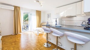 Une cuisine moderne avec des meubles blancs, un coin bar doté de tabourets blancs et une vue sur un balcon aménagé avec du mobilier d’extérieur.