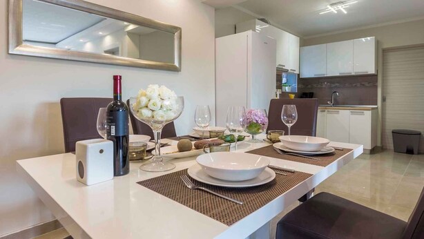A modern dining area with a white table set for two, a wine bottle, and a bowl of flowers. - Holiday home with outdoor pool (Kujići)