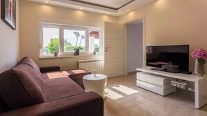 A modern living room with a brown sofa, a white coffee table, a TV on a white stand, and a window with plants. - Holiday home with outdoor pool (Kuji ći)