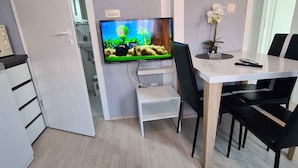 A modern living room with a flat-screen TV mounted on the wall, a dining area with a table and chairs, and a kitchen visible through an open door.