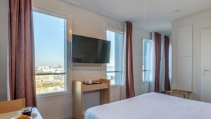 Ein Hotelzimmer mit einem Bett, einem Schreibtisch mit einer Kaffeetasse, einem an der Wand montierten Fernseher und großen Fenstern mit Vorhängen.