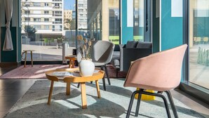 Ein modernes Hotelzimmer mit großen Fenstern, einer Couch, einem Sessel, einem kleinen Tisch und einer Vase mit Trockenblumen.
