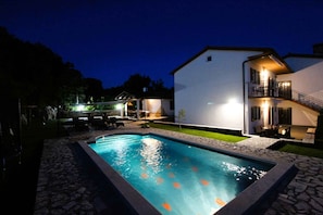 A swimming pool with a stone border, a white building with a balcony, and outdoor furniture.