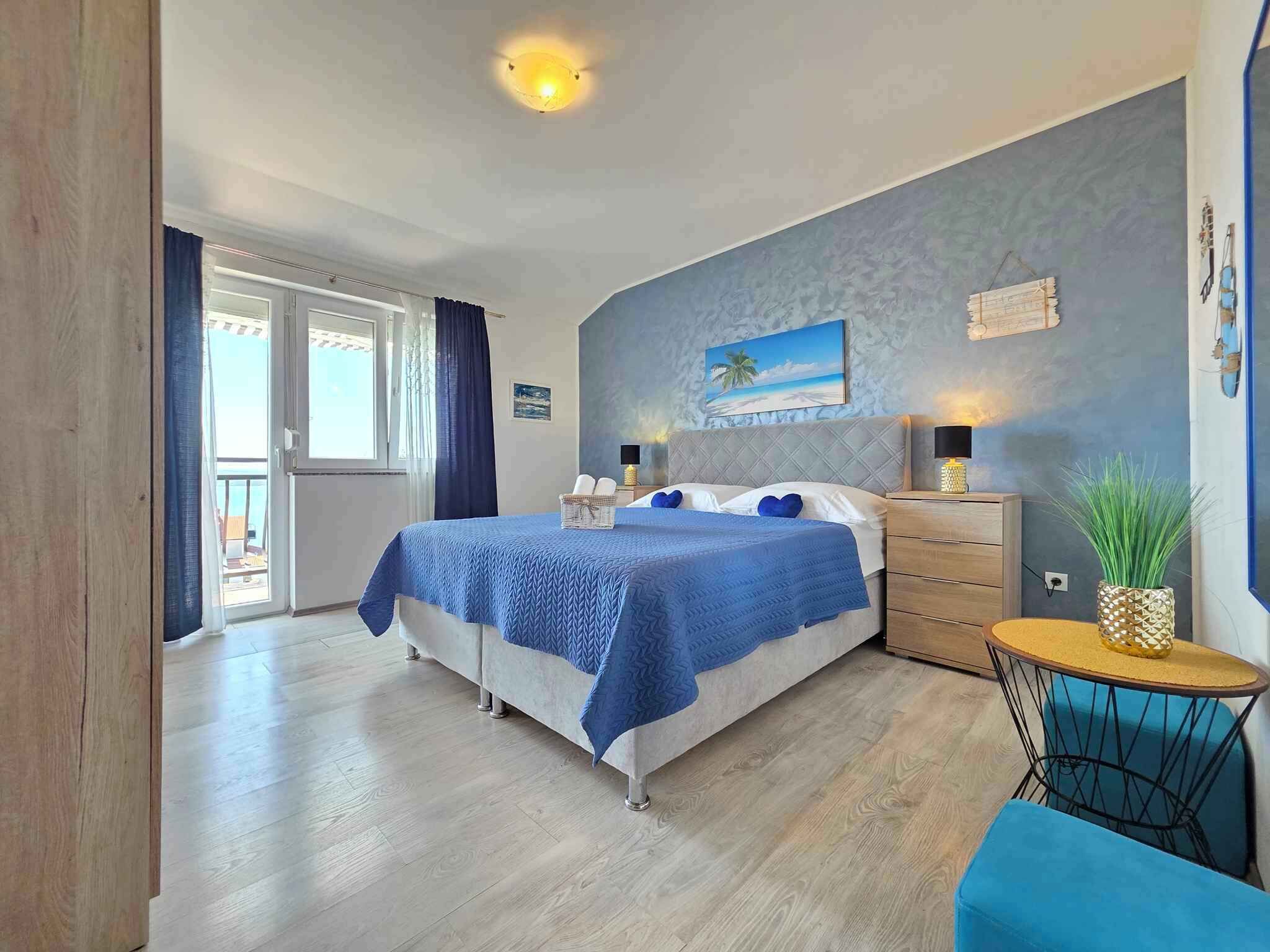 A bedroom with a bed, a nightstand, a small round table with a plant, and a view of the outside.