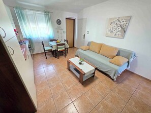 A living room with a sofa, a coffee table, a dining table with chairs, a kitchen area, and a door.