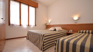 A hotel room with two beds, a wooden headboard, a window with curtains, and a wall-mounted light fixture. - Holiday apartment with balcony (Lignano)