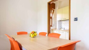 A dining area with a wooden table and orange chairs, a fruit bowl on the table, and a kitchen in the background. - Holiday apartment with air conditioning and terrace in Lignano (Lignano)