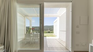 Una habitación con puerta corrediza de vidrio que da a un patio con mesa y sillas, vista a un paisaje verde y un interior blanco.
