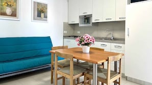 Un espacio habitable compacto con un sofá azul verdoso, una mesa de comedor de madera con cuatro sillas, una zona de cocina con armarios blancos y una pequeña maceta con flores rosadas sobre la mesa.