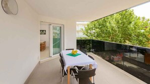 Un balcón con mesa y sillas, una puerta corrediza de vidrio y vista a árboles y edificios.