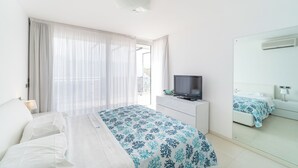 A hotel room with a bed, a TV on a dresser, a window with sheer curtains, and a large mirror.