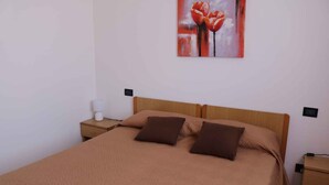 A neatly made bed with a brown quilt and two pillows, a bedside table with a lamp, and a painting of red flowers on the wall.