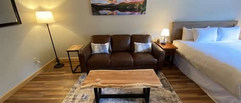 A hotel room with a brown leather sofa, a wooden coffee table, a bed with white linens, a painting of a mountain landscape, and two bedside lamps.