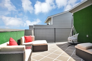 Une terrasse extérieure aménagée avec des meubles en osier, un fauteuil suspendu et une vue sur un bâtiment doté d’un mur végétal et d’une clôture grise.