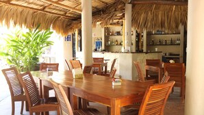 A wooden dining table with chairs, a bar area with shelves of bottles, and a large plant in the foreground. - Hotel Circulo Bacalar (Bacalar)