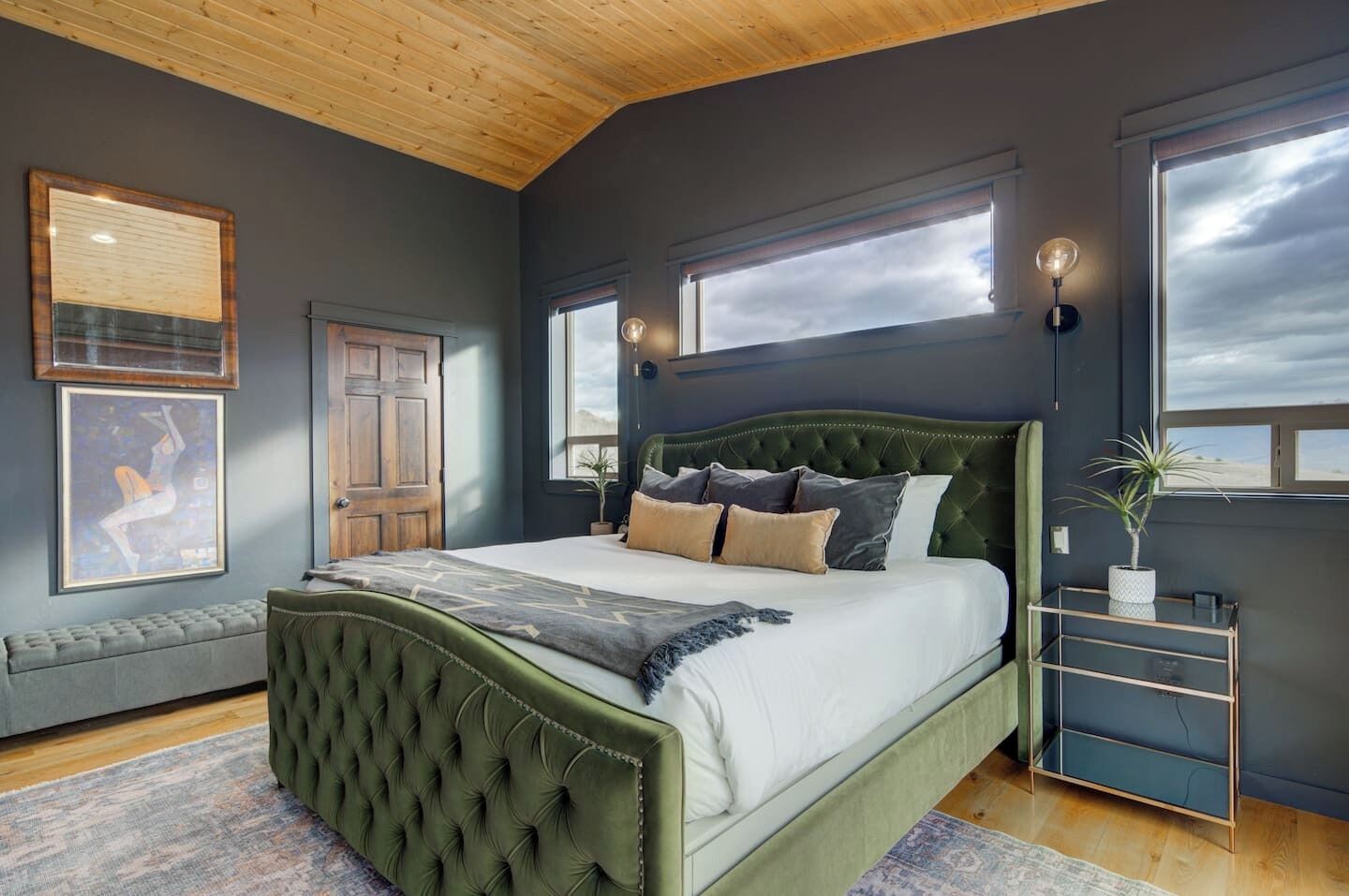 A bedroom with a green upholstered bed, a bench, a bedside table, and a large window.