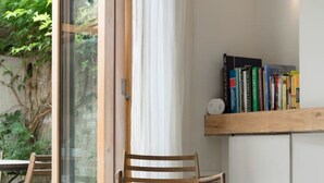 Une table en bois avec une bougie et une sculpture en pierre, une chaise, une bibliothèque remplie de livres et une porte en verre donnant sur un espace extérieur verdoyant.